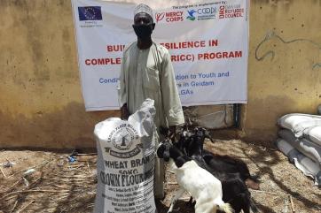 A smallholder farmer poses with goods and livestock received through the bricc program.