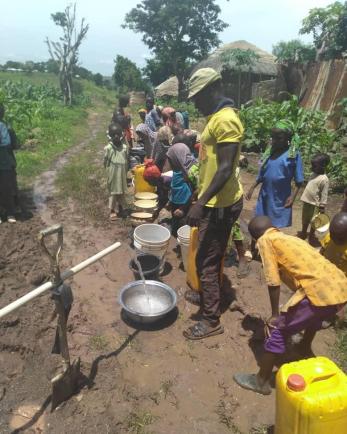 People stand around water coming from a hand pump borehole in nigeria.