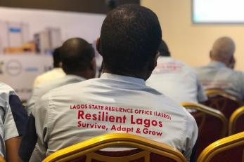 A person sits in a chair at a presentation wearing a shirt that reads lagos state resilience office, resilient lagos, survive, adapt, and grow.