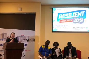 A person at a podium holds a microphone while three people sit nearby in chairs under a screen with logo for a special event called resilient lagos week 2019.