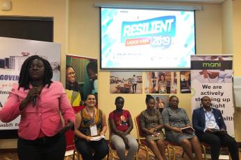 A person speaking into a microphone leads stands in front of a panel of people sitting in chairs under a projection screen displaying the words Resilient Lagos Week 2019.