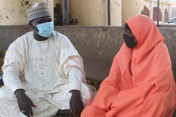 Two people sit next to each other and exchange glances while wearing cloth face masks.
