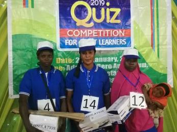 Three young people wearing numbered bibs stand in front of a competition poster holding consolation prizes. 