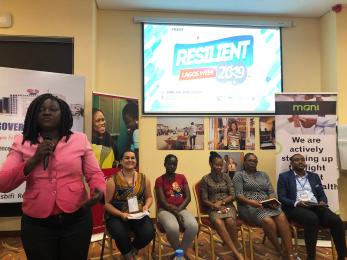 A person speaking into a microphone leads stands in front of a panel of people sitting in chairs under a projection screen displaying the words resilient lagos week 2019.