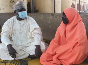 Two people sit next to each other and exchange glances while wearing cloth face masks.