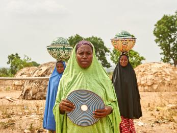 Three nigerian woman in traditional dress; two with balanced loads on their heads.